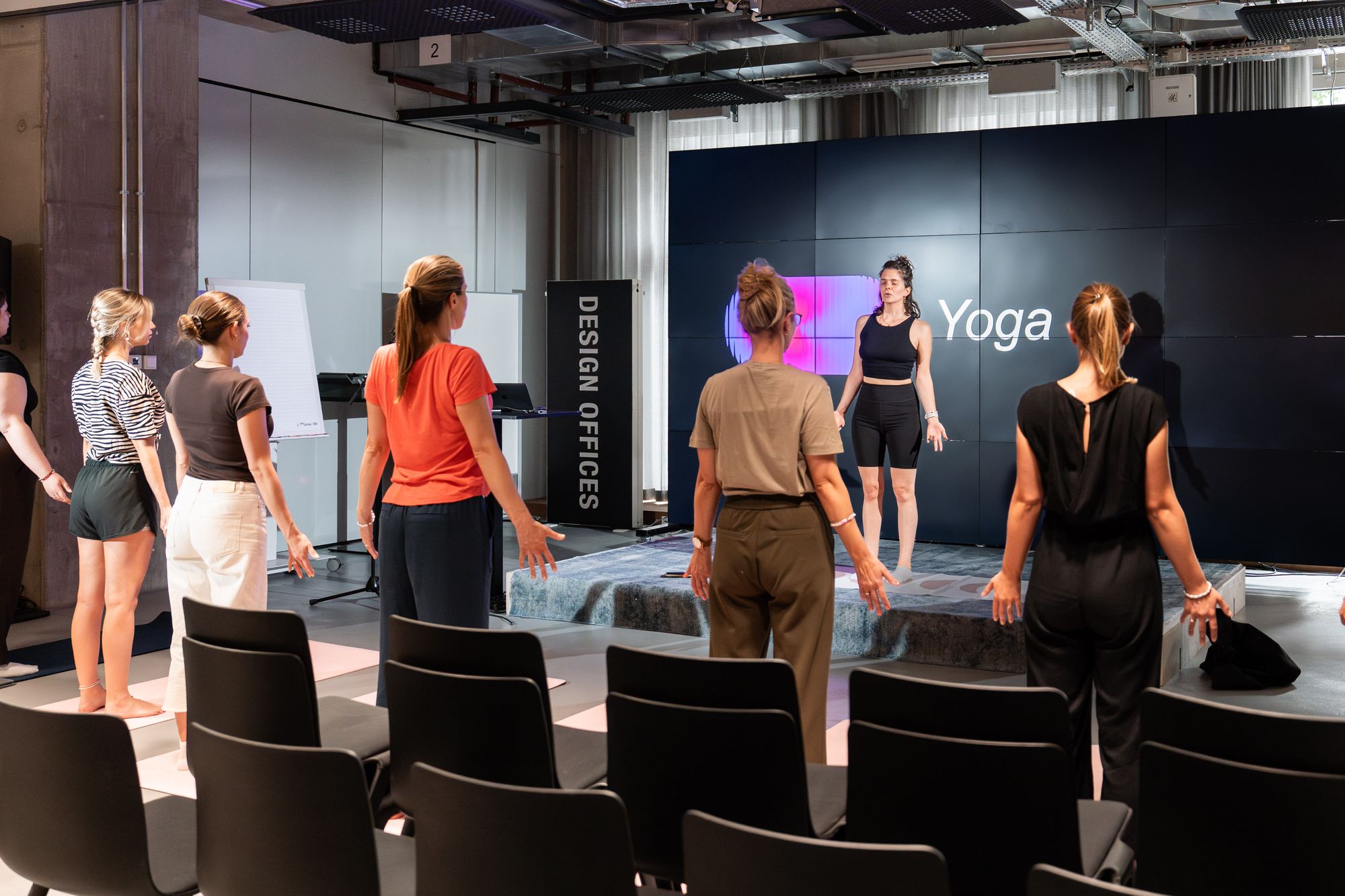 Eine Yogalehrerin leitet eine Klasse mit sechs Frauen in einem modernen Studio mit einem „Yoga“-Schild an der Wand.