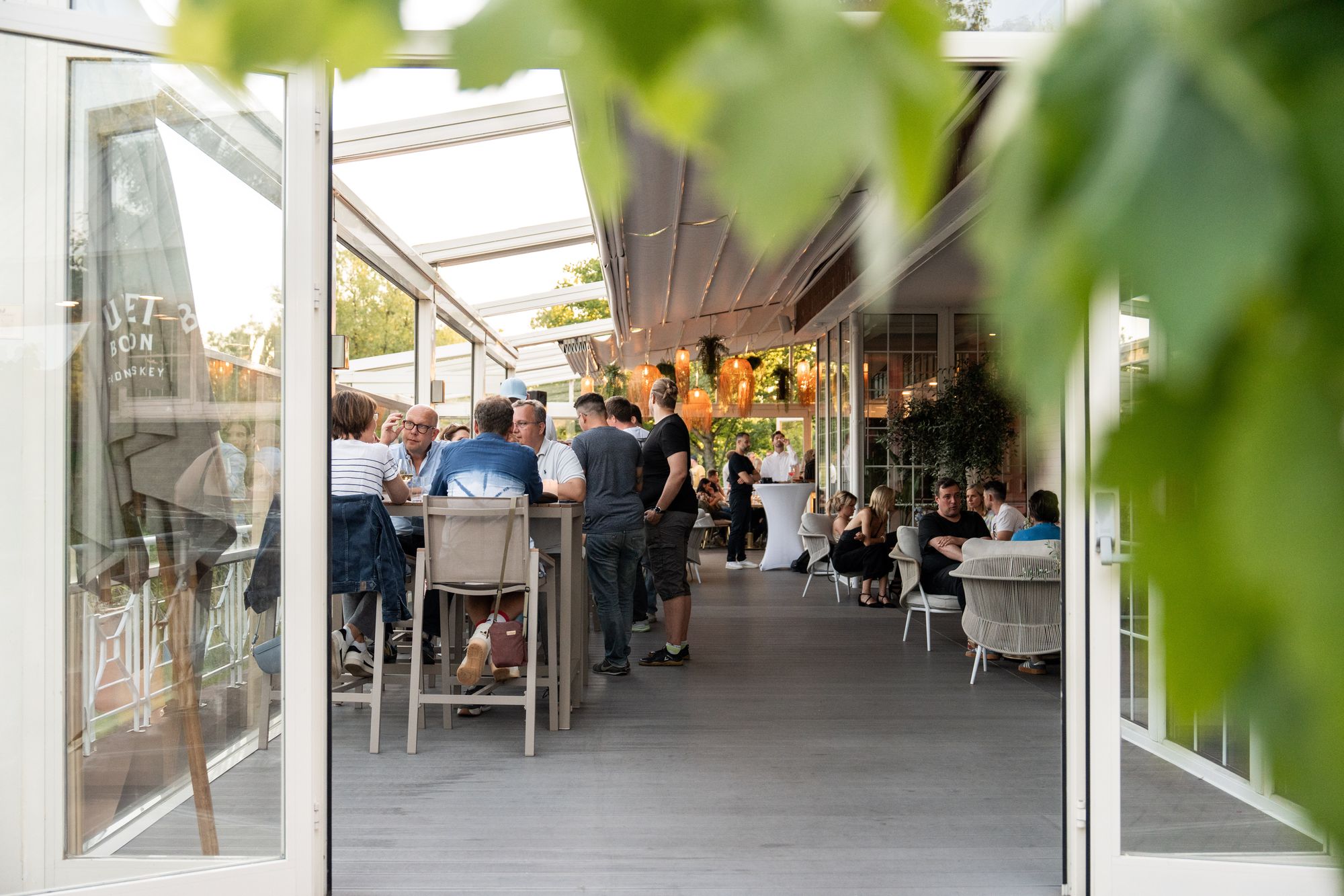 Menschen unterhalten sich in einem Freiluftrestaurant mit offenen Türen und viel Grün unter einer teilweise überdachten Terrasse.