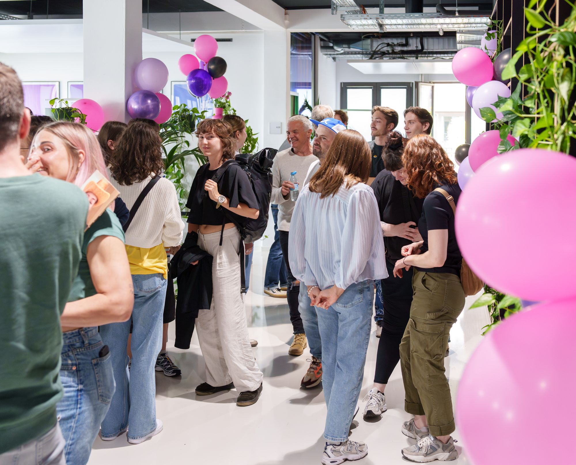 A group of people socializing in a modern interone office space decorated with pink and purple balloons and greenery.