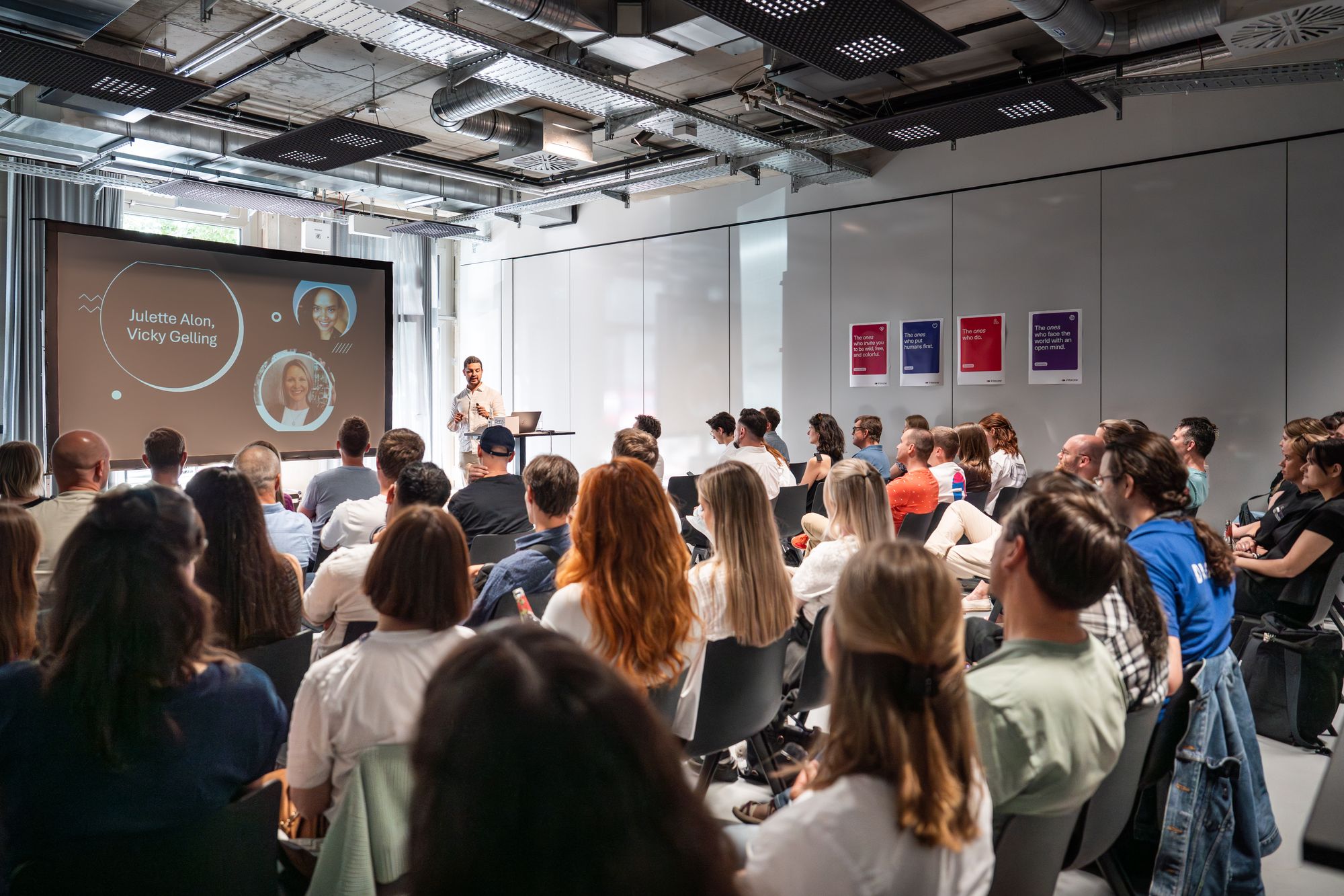 Ein Redner präsentiert einem sitzenden Publikum in einem modernen Konferenzraum mit einem großen Bildschirm, auf dem Namen und Fotos angezeigt werden.