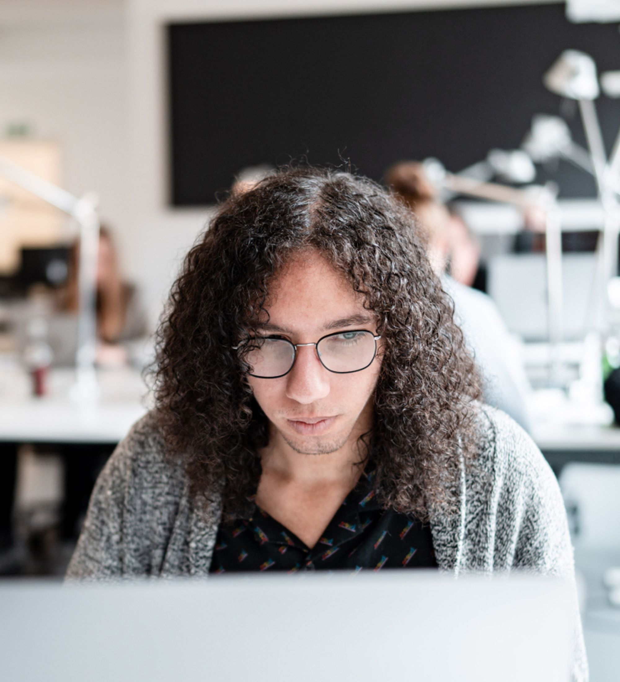 Person mit Laptop arbeitet konzentriert in einem modernen, offenen Büro mit Team im Hintergrund.