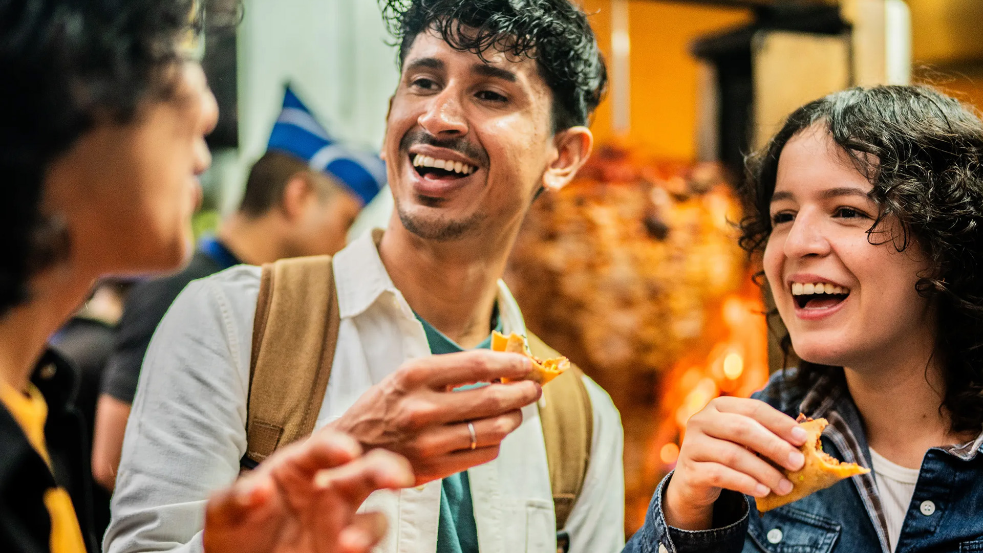 Three people smiling and eating snacks together indoors, with a warm and lively background ambiance.