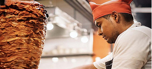 A cook wearing a red hat prepares a large vertical spit of meat in a kitchen.