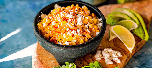 Bowl of Mexican street corn topped with cheese, chili powder, and lime wedges on a wooden board.