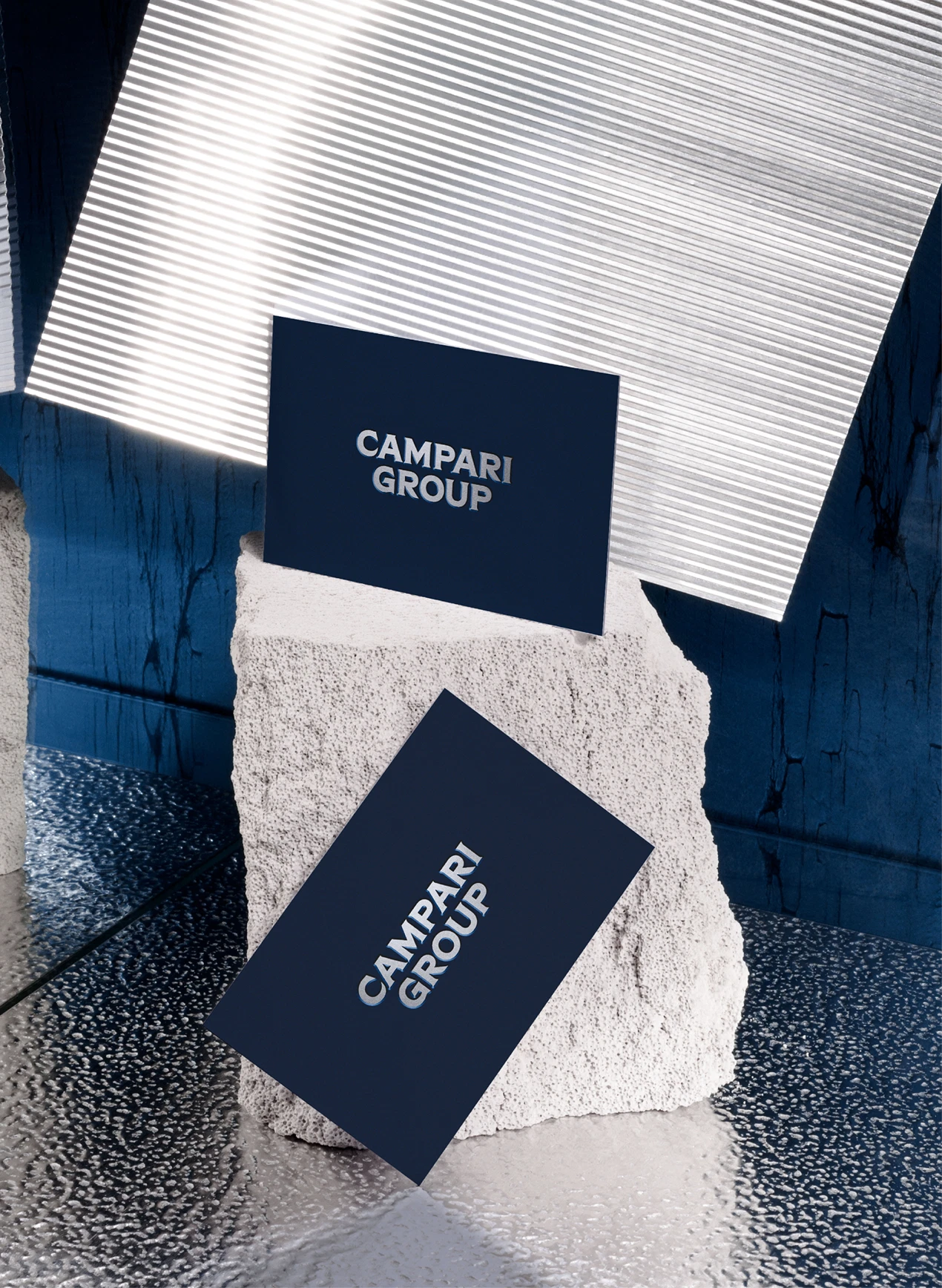 Two dark blue cards with "CAMPARI GROUP" text, positioned on and against a textured stone block, with metallic and blue surfaces around.