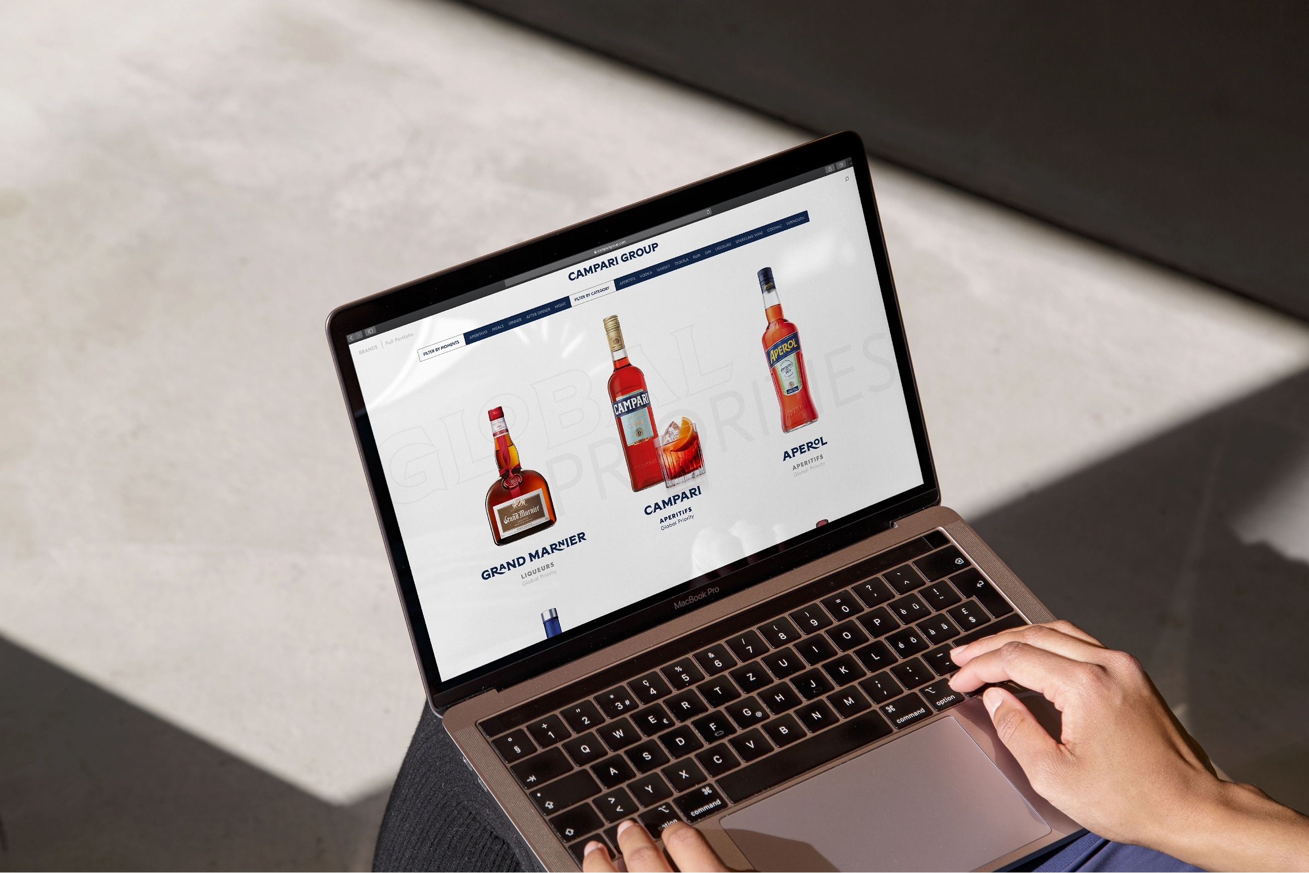 Person using a laptop displaying a webpage with images of Grand Marnier, Campari, and Aperol bottles from the Campari Group website.