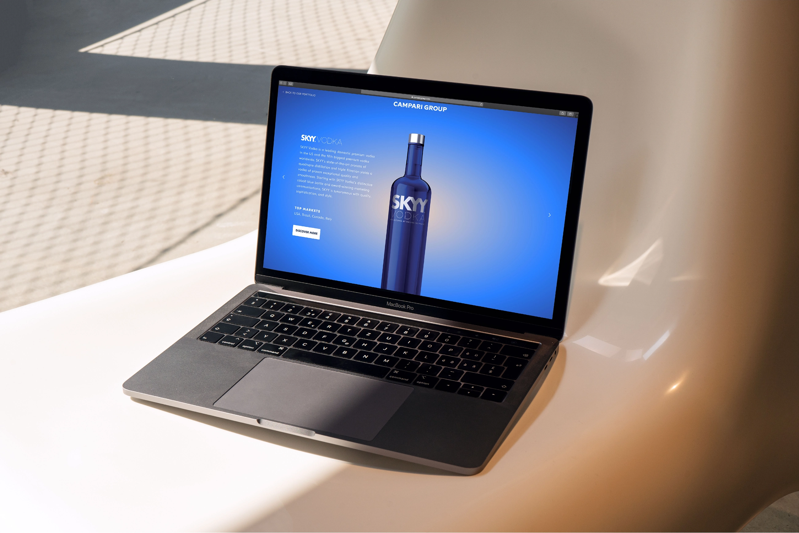 A laptop displays a webpage featuring SKYY Vodka, with a blue interface, placed on a sleek white surface under soft lighting.
