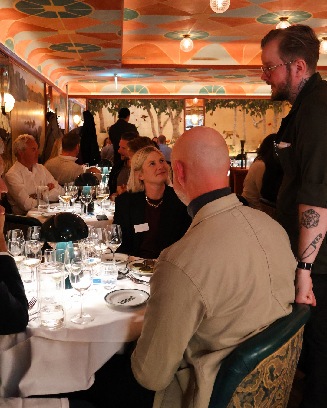 Guests dining at an elegant restaurant with an orange-patterned ceiling, wine glasses on white tablecloths, and a server chatting with the guests at the tables.