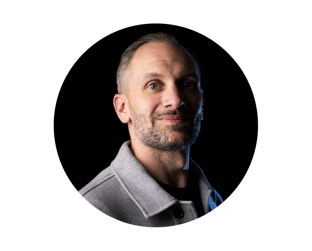 Man with short hair and beard in a grey jacket, smiling slightly against a black background.