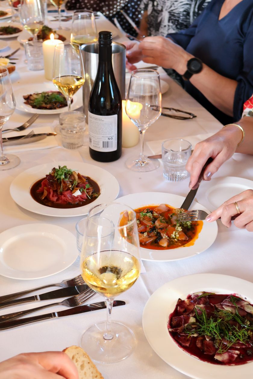 Elegant dinner table with wine glasses, a bottle of white wine, candles, and colorful plated dishes, with diners eating.