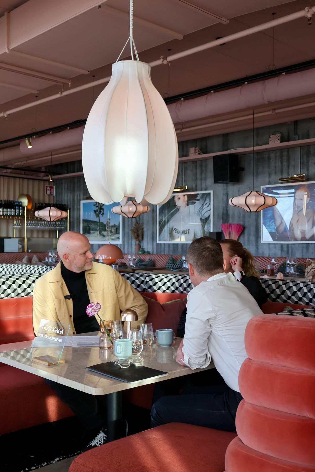 Three people having dinner at an elegant restaurant with booths upholstered in pink velvet, chandeliers, and an eclectic interior featuring artwork on the walls.