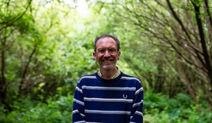 Man in blue striped jumper, wearing glasses, smiling at the camera in a forest