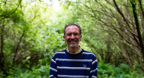 Man in blue striped jumper, wearing glasses, smiling at the camera in a forest