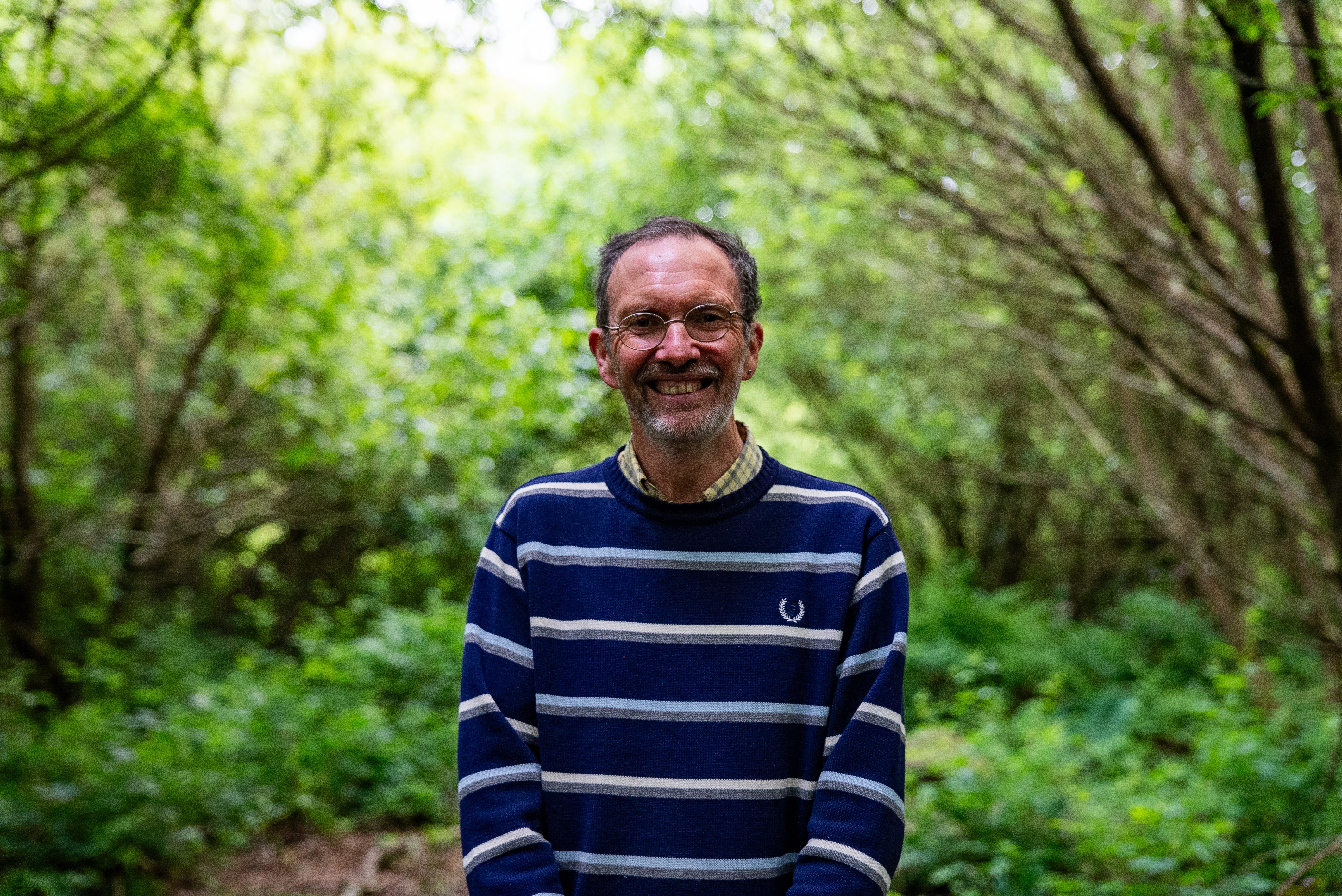 Man in blue striped jumper, wearing glasses, smiling at the camera in a forest