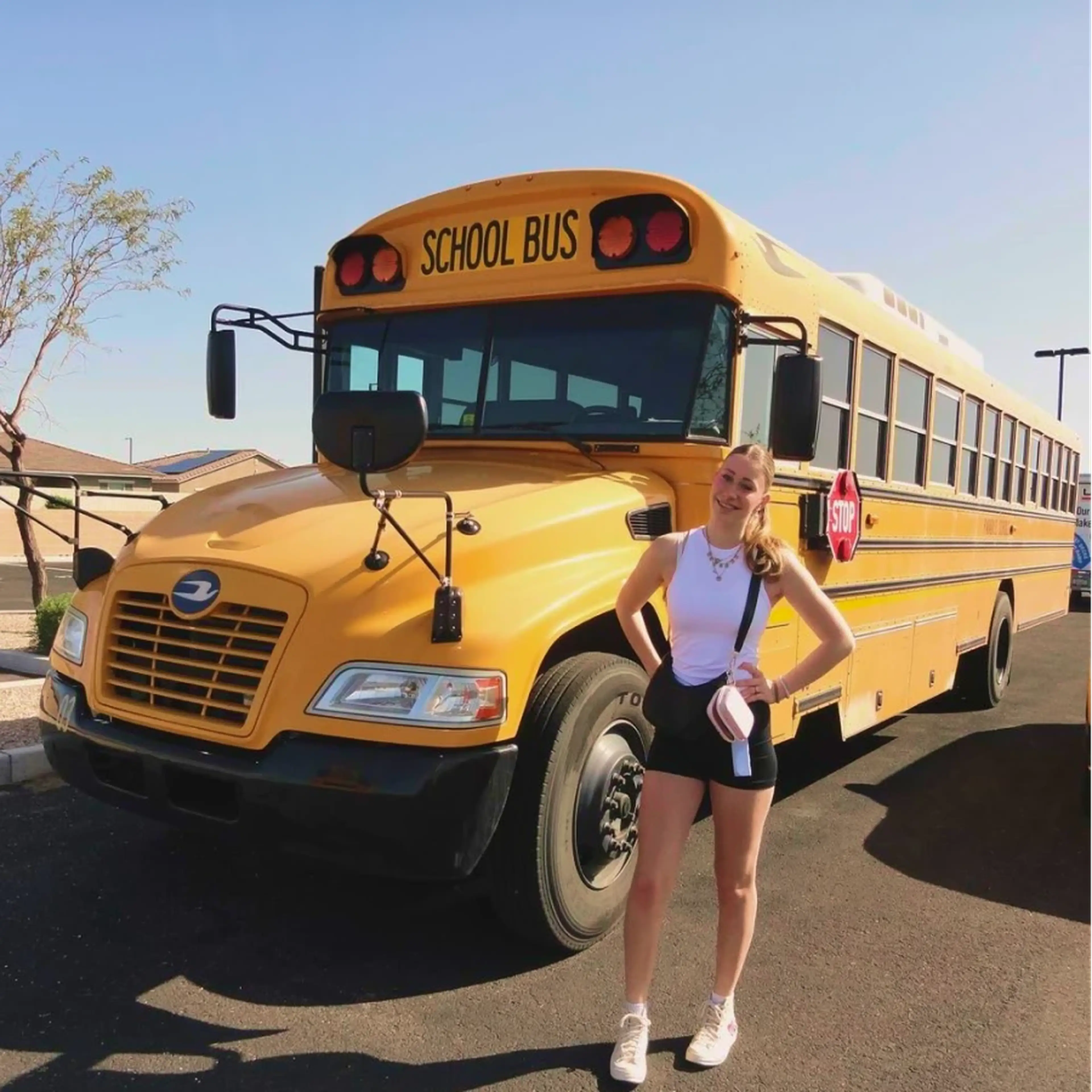 Smiling person posing in front of a parked yellow school bus on a sunny day.