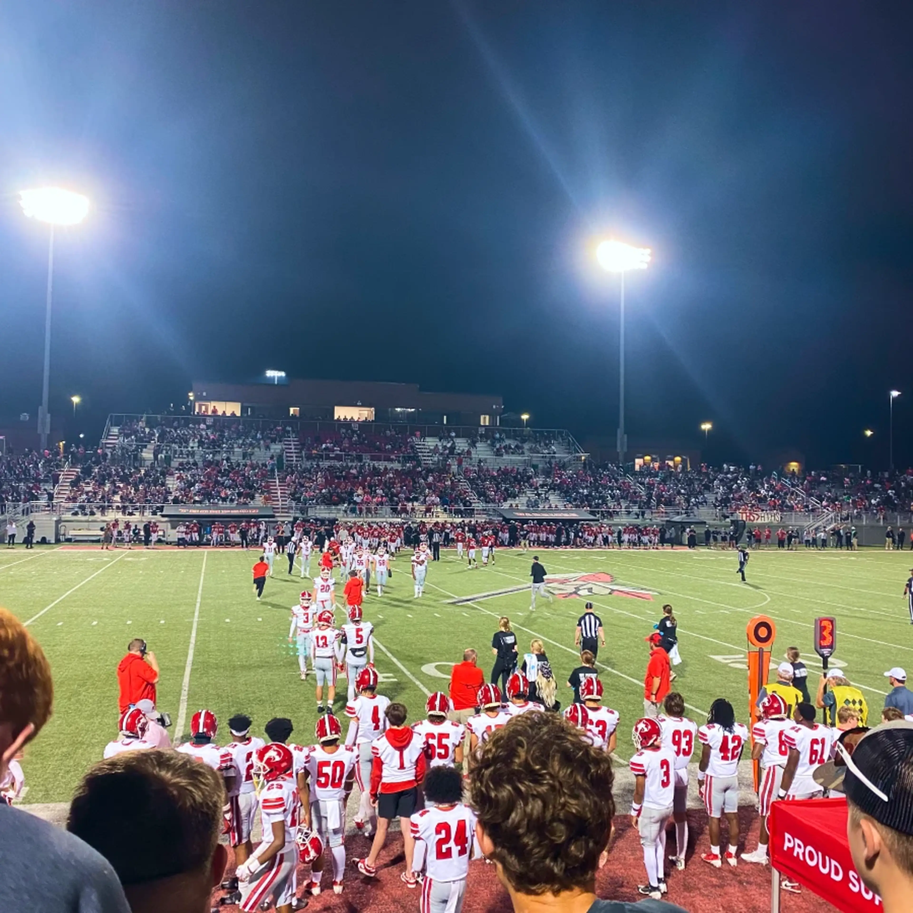 Nighttime high school football game, players in red and white gear on the field, with full bleachers and bright stadium lights.