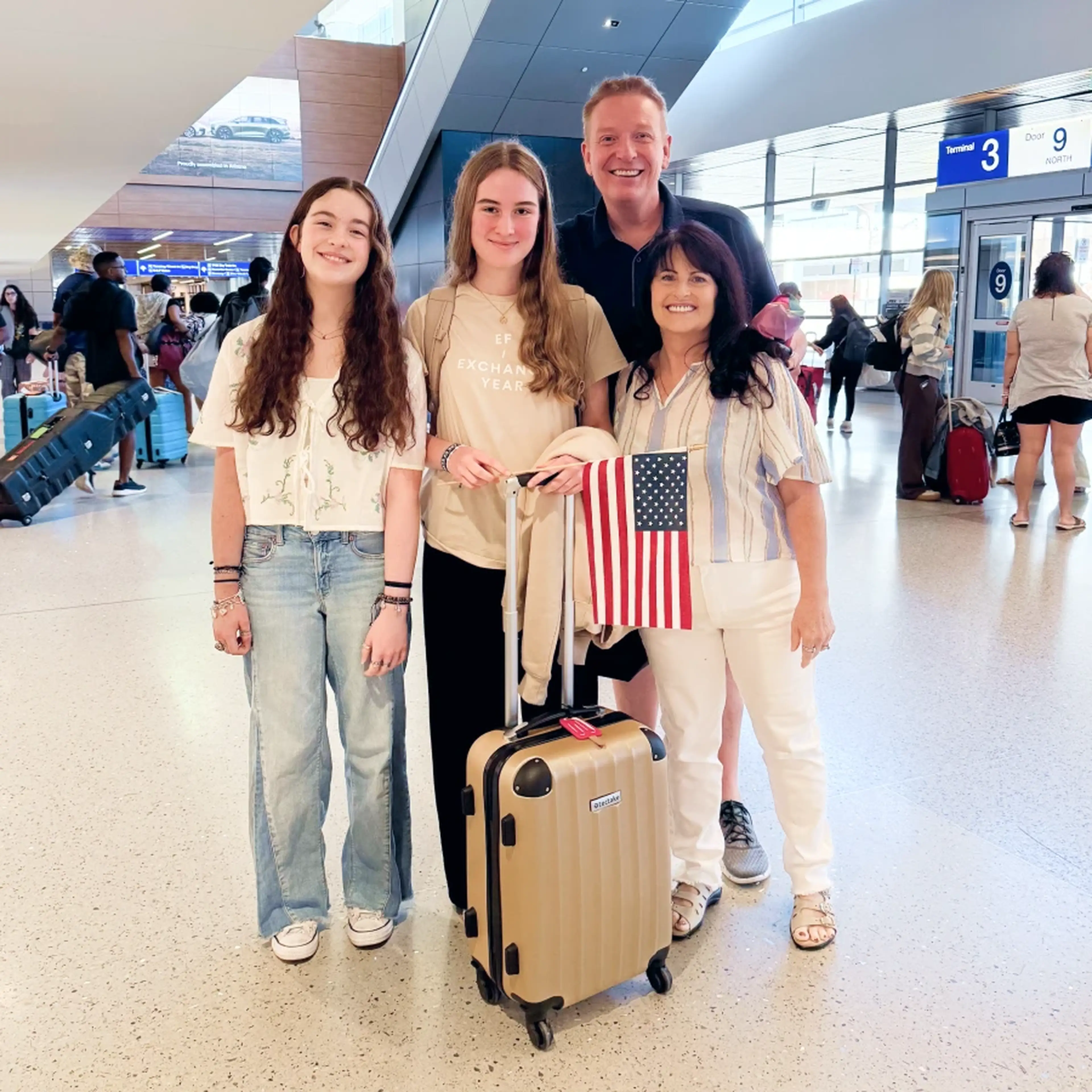 A smiling group of four people at an airport, with a suitcase and an American flag.