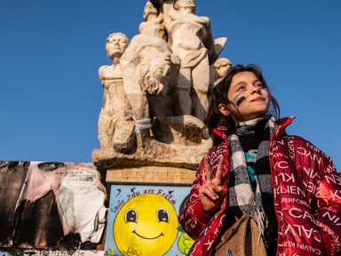 Ein junges Mädchen posiert vor einer zerstörten Statue und einem abgebrannten Transparent von Bashar al-Assad.
