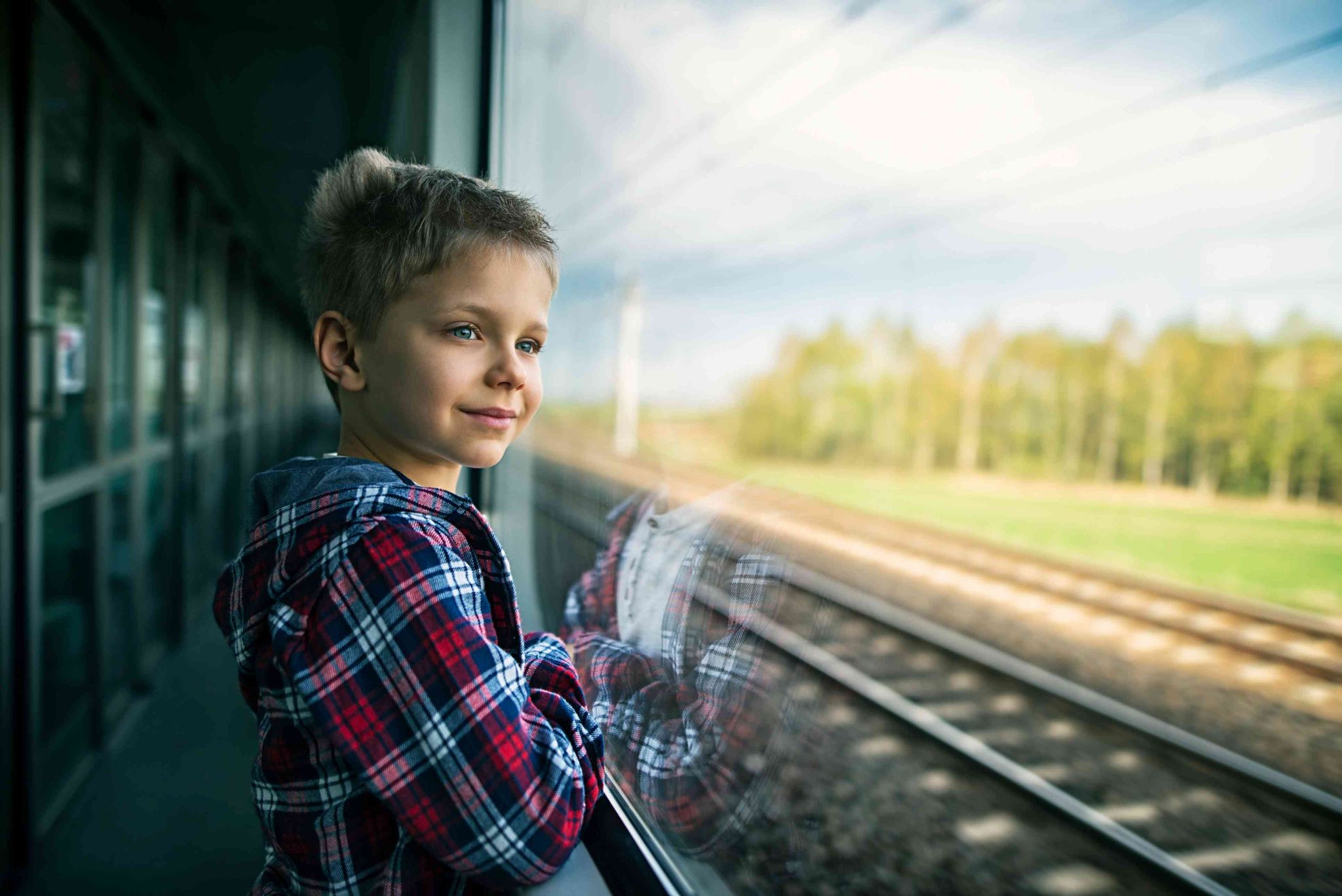 Pojke i rutig skjorta tittar ut genom ett tågfönster på landskap med träd och järnvägsspår.
