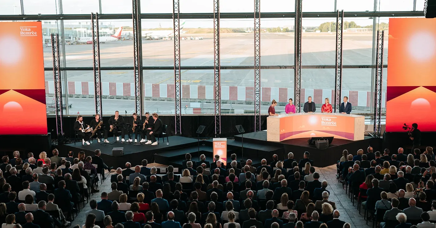 An event at an airport terminal with a large audience, a panel of speakers, and musicians, featuring large displays with "Voka Rentree."