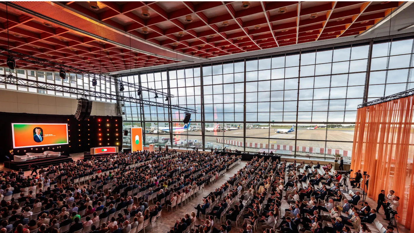 Large audience attending a conference in a spacious hall with a view of the airport runway and parked planes through large windows.