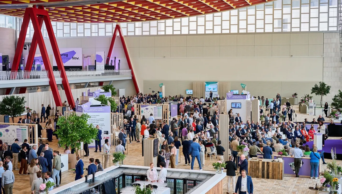 A bustling conference with attendees gathered around booths and stages in a bright, modern venue with large red beams and greenery.