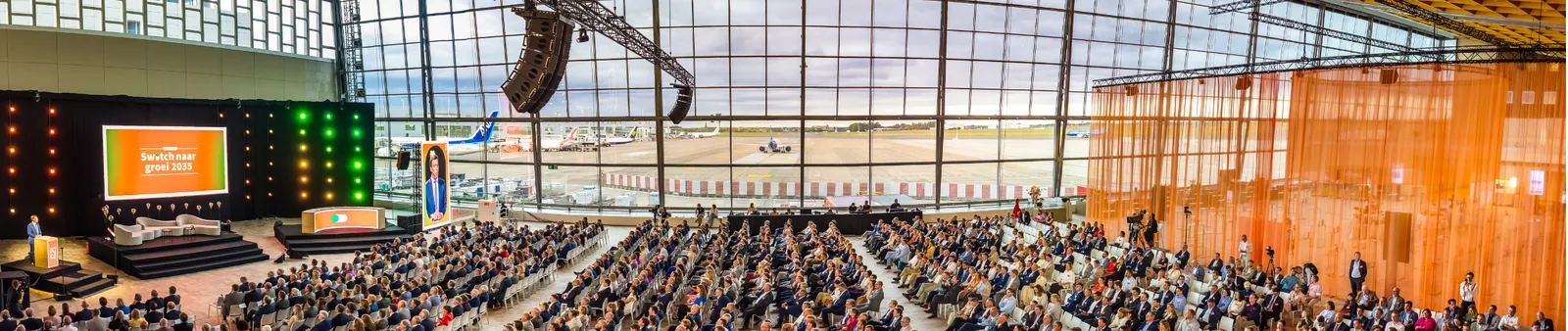 A large audience in a spacious conference hall overlooks an airport runway, with speakers on stage beside screens and a city view in the background.