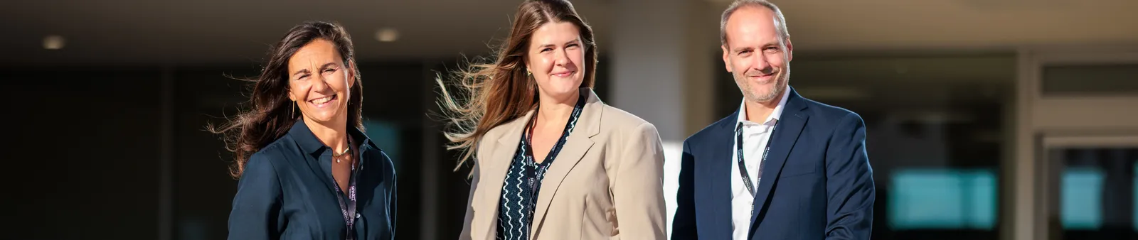 Three people in professional attire standing outside, smiling warmly at the camera.