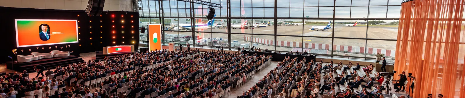 An audience at a large airport conference with planes visible through massive windows. A presentation is displayed on a large screen.