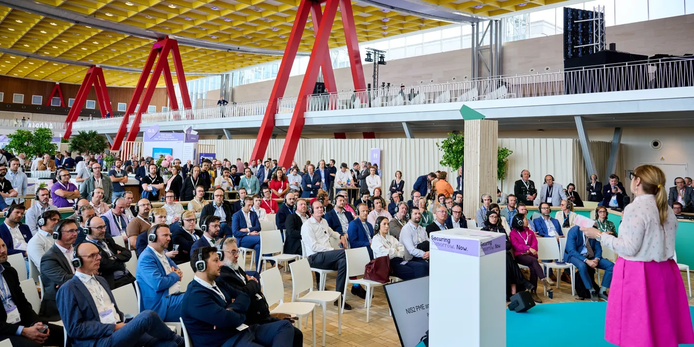 A speaker addresses a large, attentive audience wearing headphones in a modern conference hall with bold red architectural features.