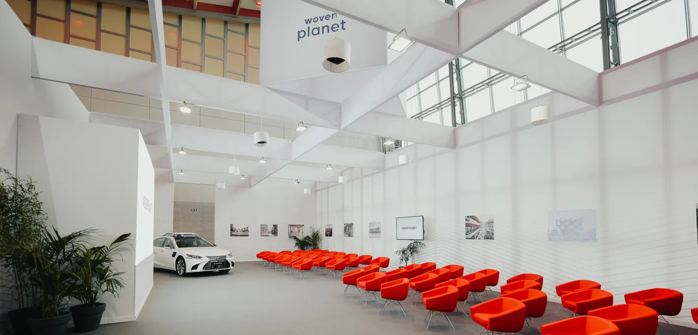 A modern exhibition space with bright red chairs, white car, plants, and the "Woven Planet" logo on the wall.