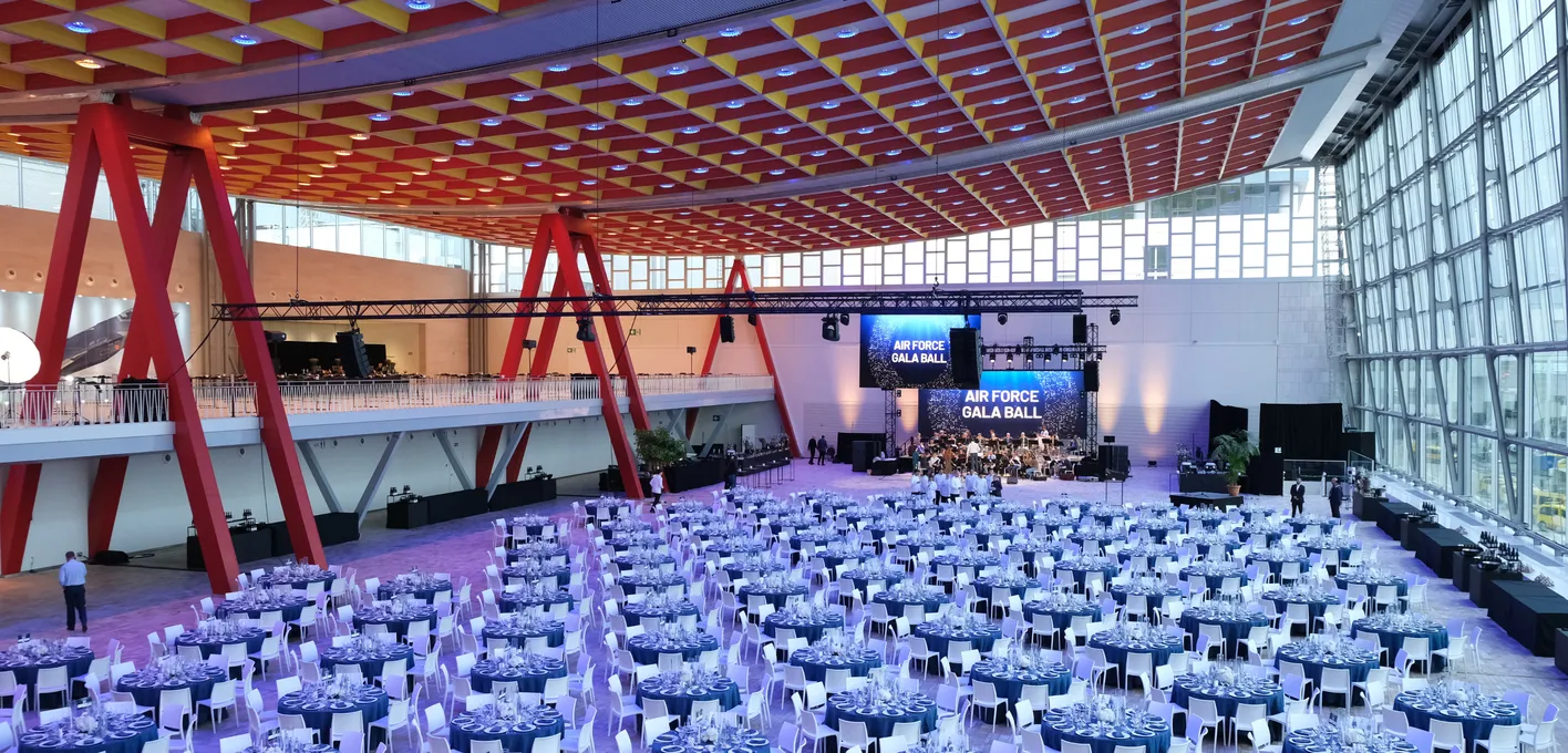 A large hall set for the Air Force Gala Ball with round tables, blue tablecloths, and a stage with lighting and screens.
