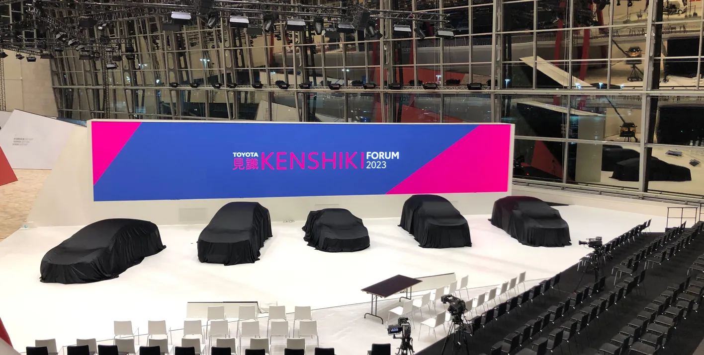 Covered cars at a display with "Toyota Kenshiki Forum 2023" on a large screen in a modern exhibition space.