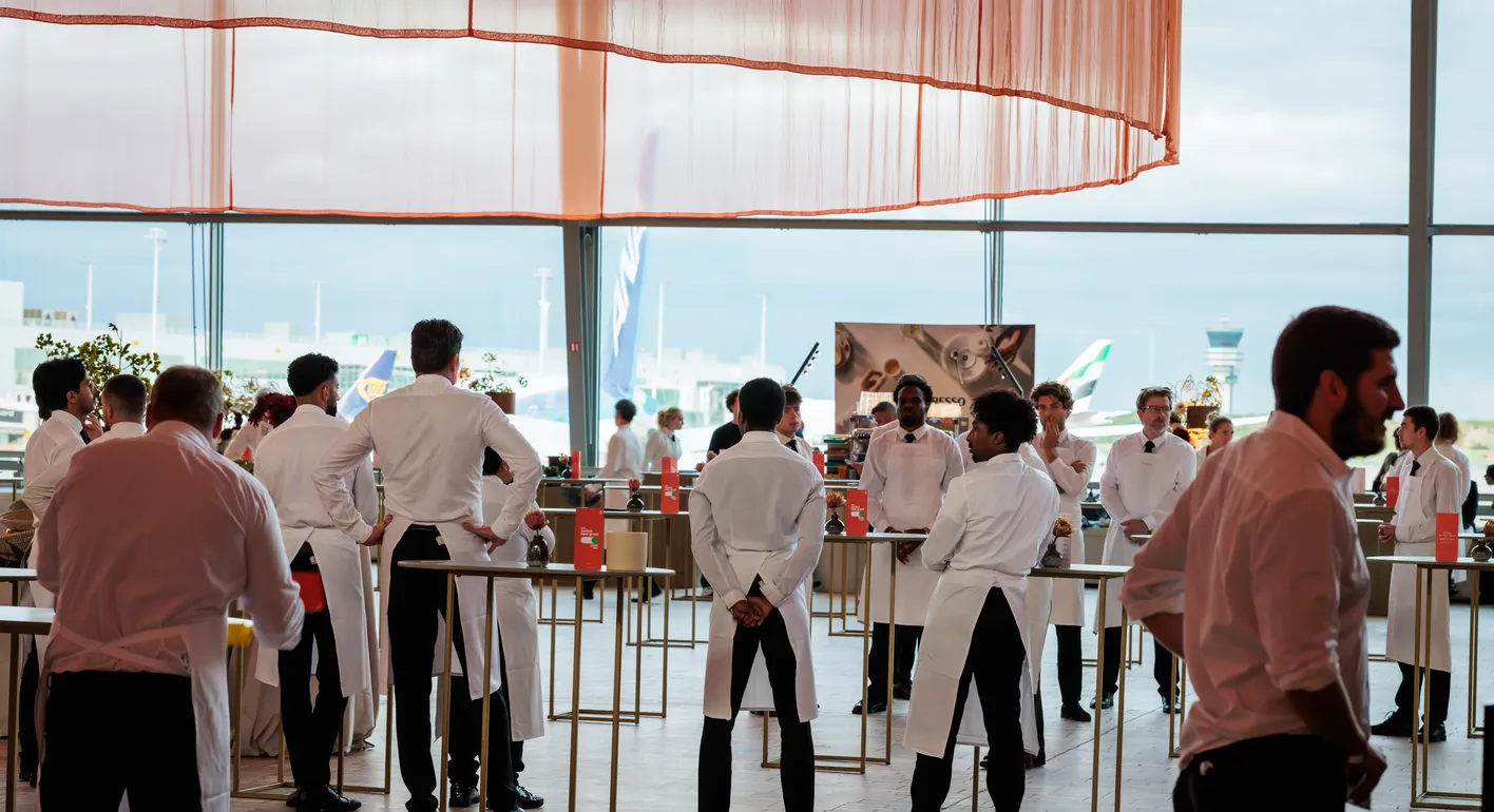 A group of servers in white aprons stand in a spacious, sunlit room with a view of an airport runway and airplanes outside.