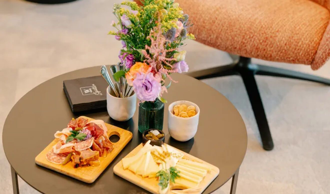 Round black coffee table with charcuterie boards (meats and cheeses), bowls of crackers and pickles, a vase of flowers and utensils.