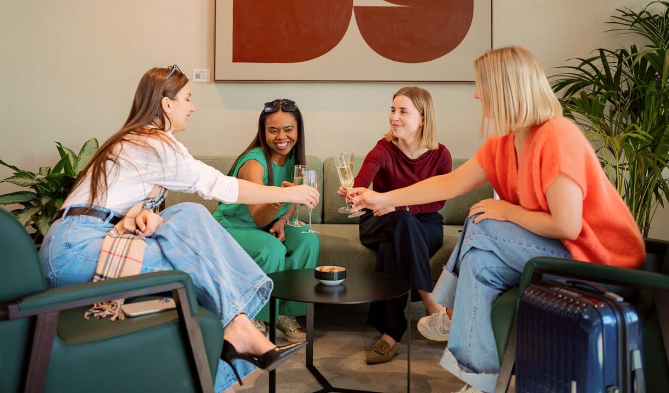 Four women seated in a lounge, smiling and toasting with champagne glasses around a small table, plants and a suitcase nearby.