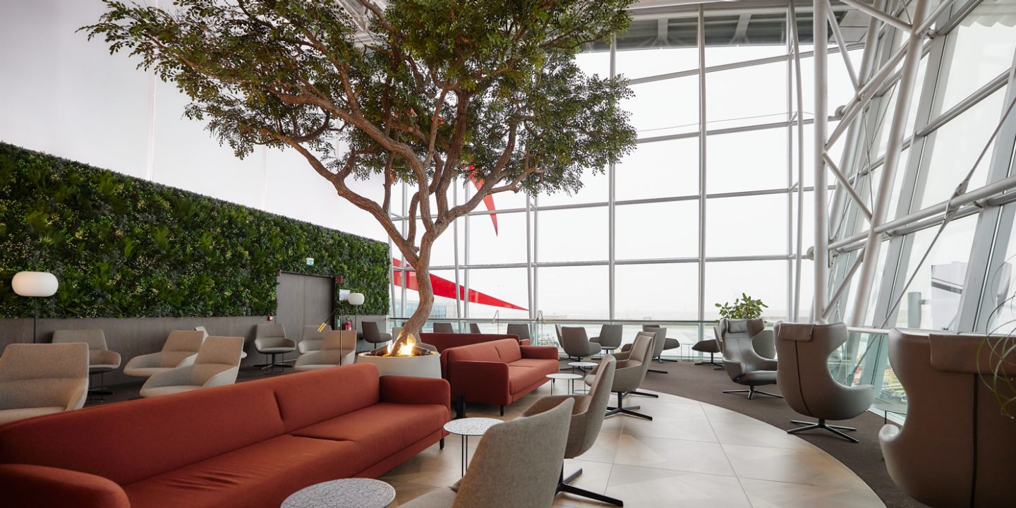 Modern airport lounge with large windows, a central tree, red couches, gray chairs, plant wall, and round tables, creating a spacious and comfortable atmosphere.