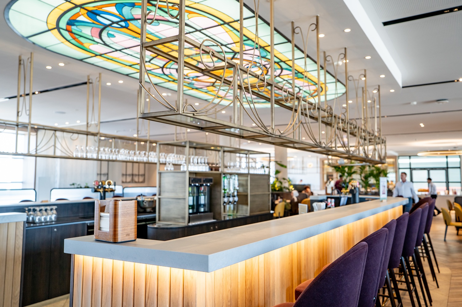 Modern bar interior with a long counter, dark purple barstools, stained glass ceiling, wine glasses, and people in the background.