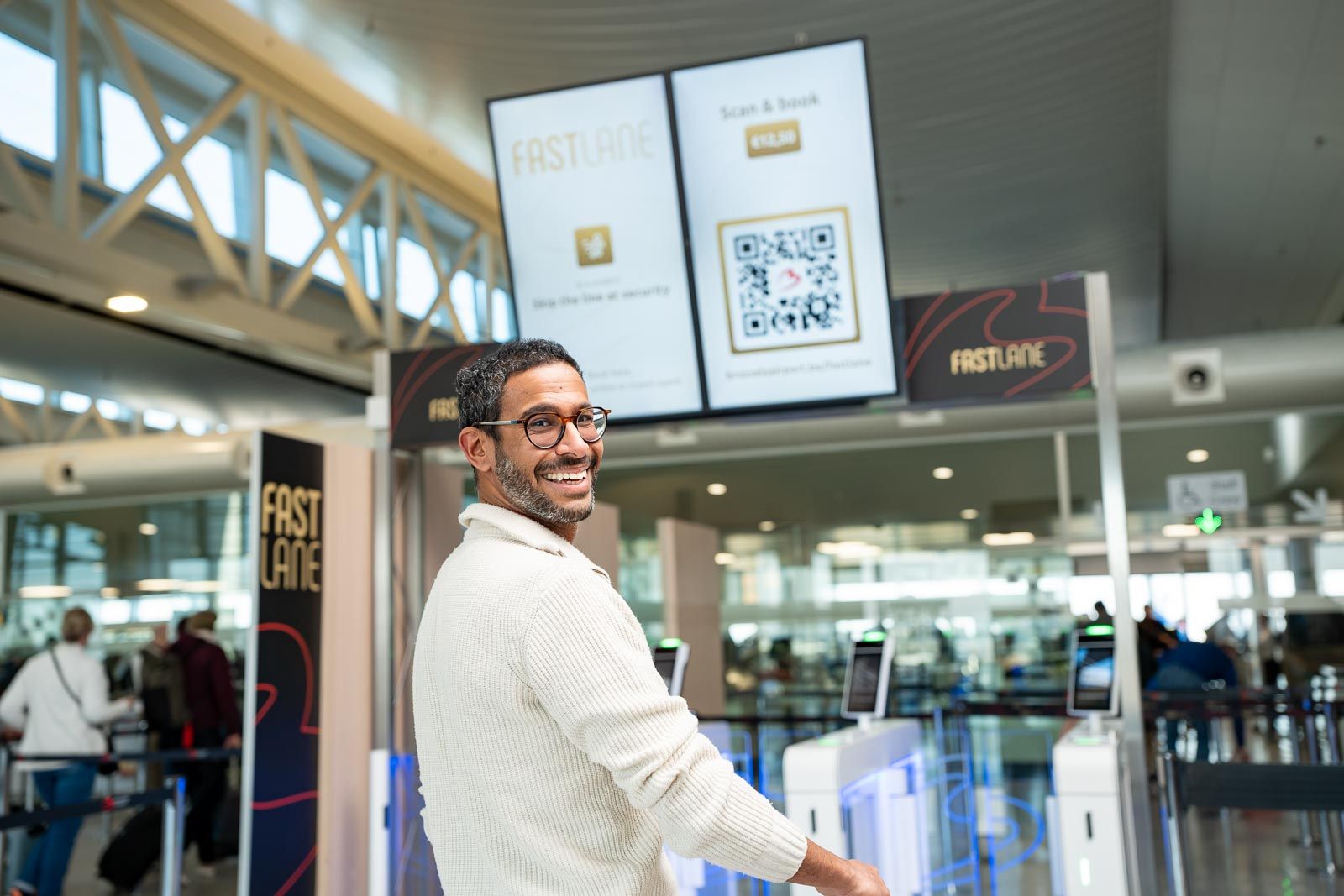 Man smiling and walking through airport security with automated gates, digital screens displaying "FASTLANE" and a QR code in the background.