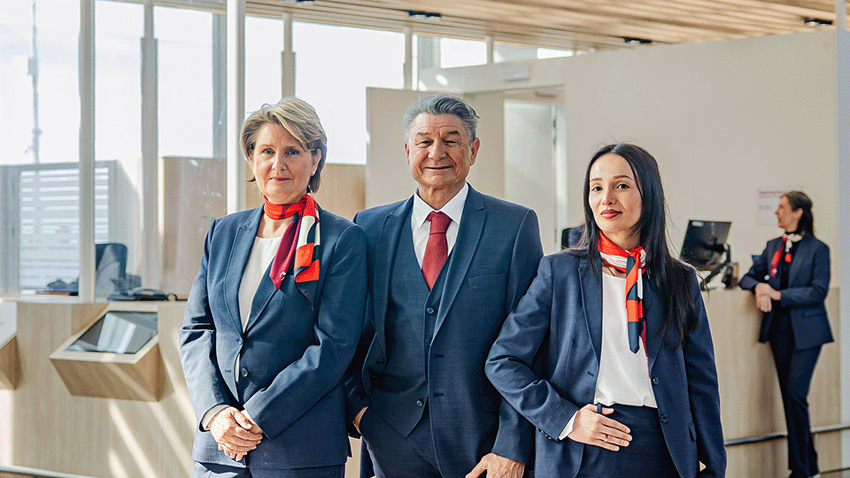 Three uniformed staff stand smiling at an information desk with a sign above them, in a modern, well-lit environment.