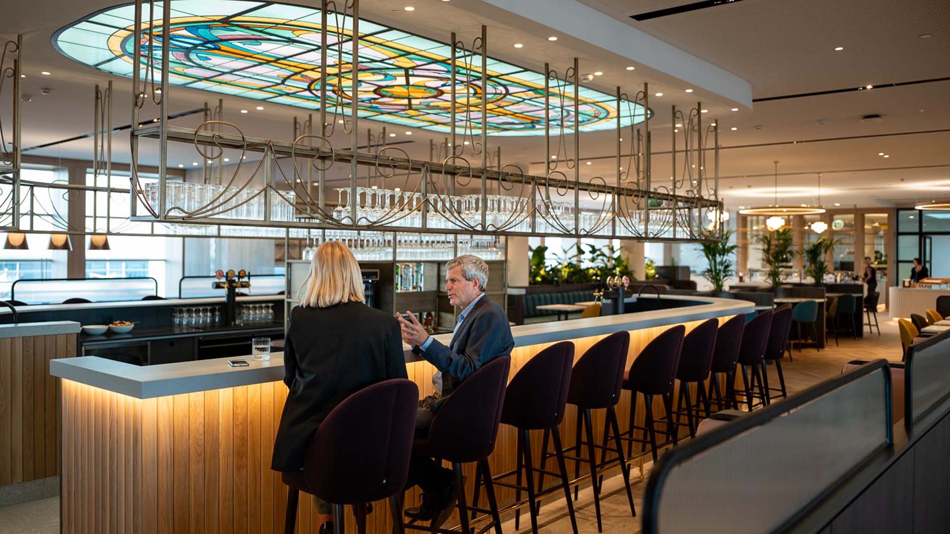 Two people sit at a modern, well-lit bar with a stained glass ceiling. The space features wooden accents and surrounding greenery.