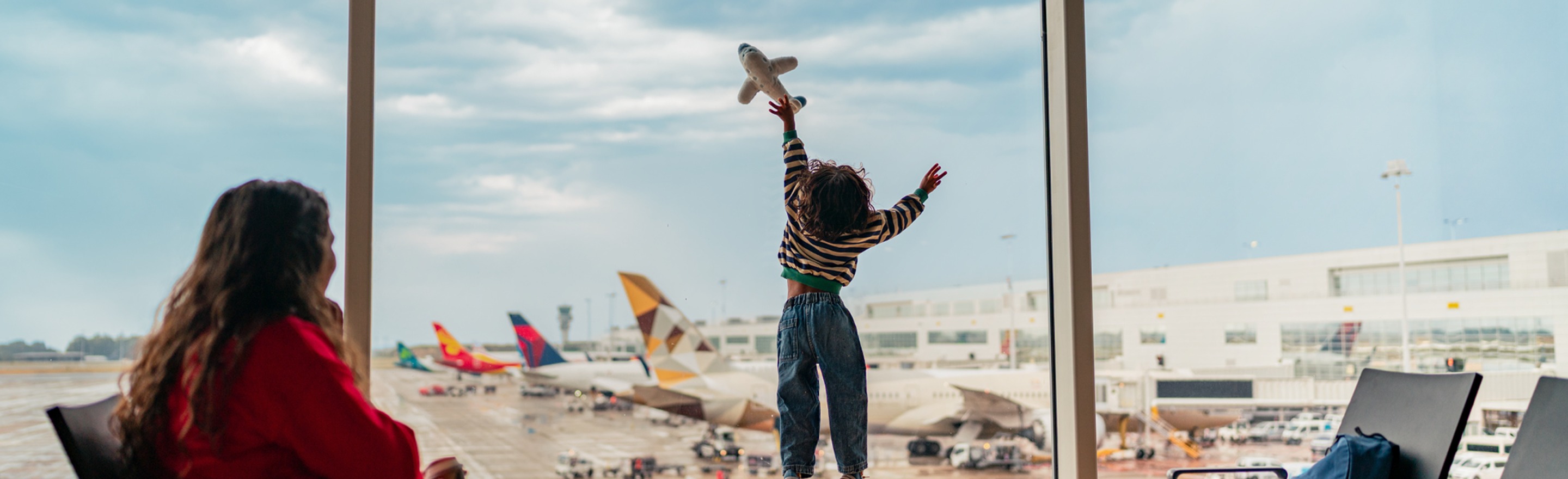 Child in a striped shirt reaches with a toy plane at airport window, overlooking parked airplanes. Adult in red observes.
