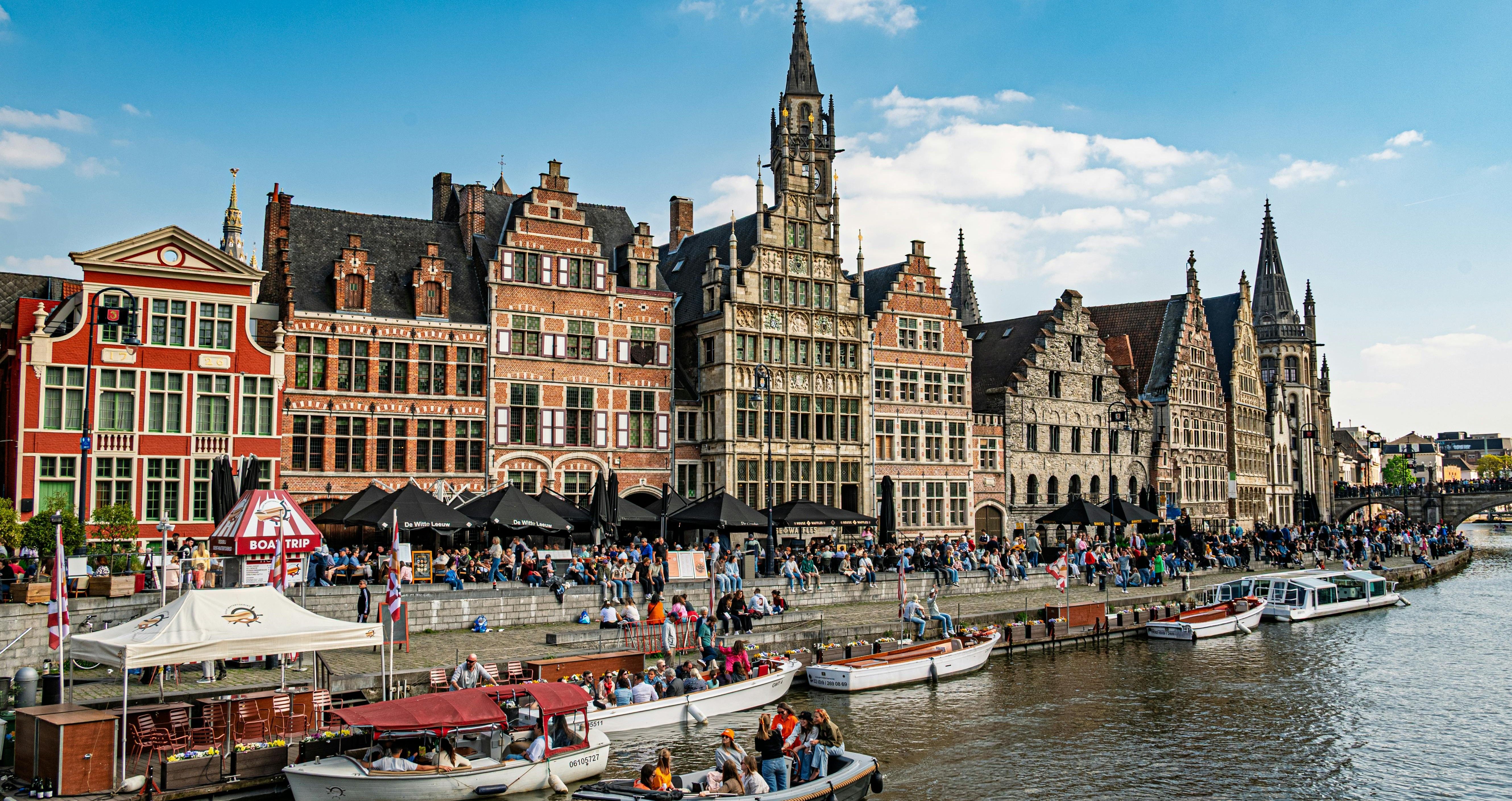 People enjoying a sunny day by the canal in Ghent, Belgium, with historic gabled buildings and boats lining the waterfront.