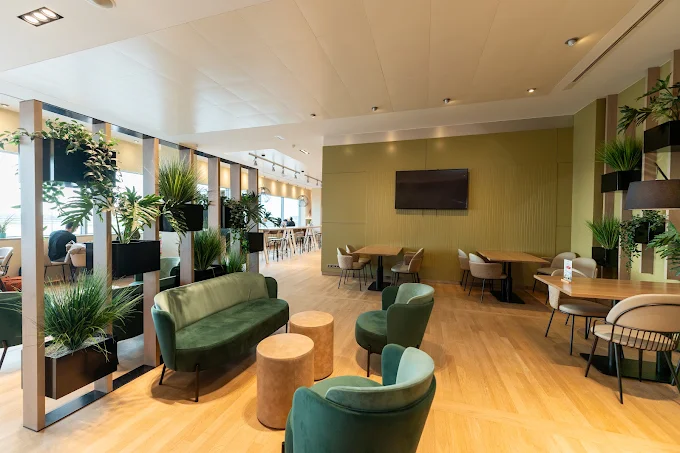Modern lounge with green sofas, wooden tables, and abundant plants. Large TV on the wall, bright lighting, and natural light from windows.