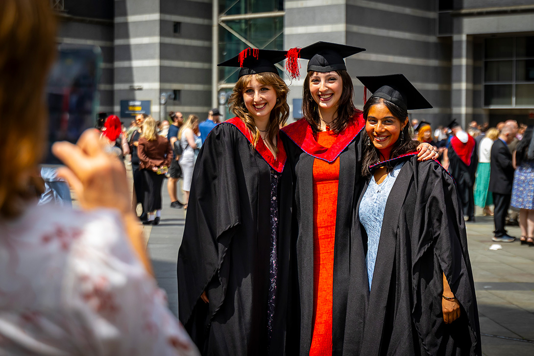 Three graduates in caps and gowns pose for a photo outdoors, smiling. Others in graduation attire are in the background.