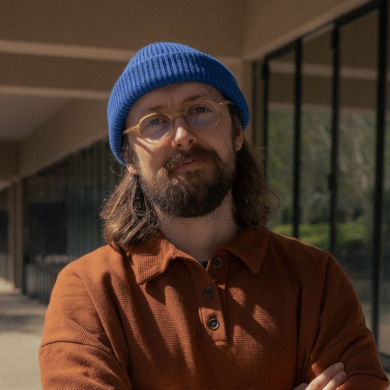 A person with shoulder-length hair and a beard wearing a blue beanie and glasses, dressed in an orange shirt, standing outside near glass windows.