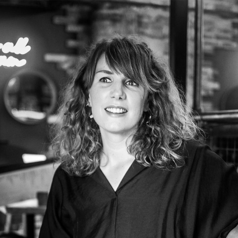 Woman with curly hair smiling, wearing a dark top, indoors. Blurred background with circular mirrors and a neon sign. Black and white image.