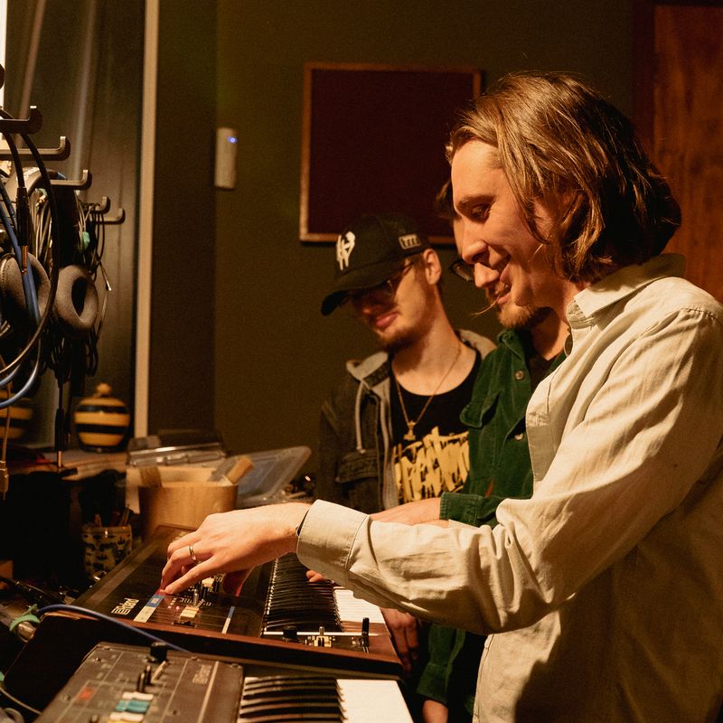 Two musicians in a studio, one playing a keyboard synthesizer while the other watches. Sound equipment surrounds them.