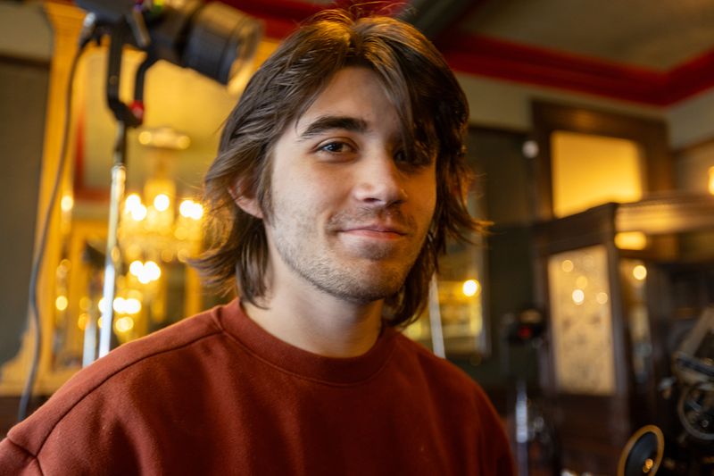 A person with long hair and a smile, wearing a red sweater, stands indoors with blurred warm lighting in the background.