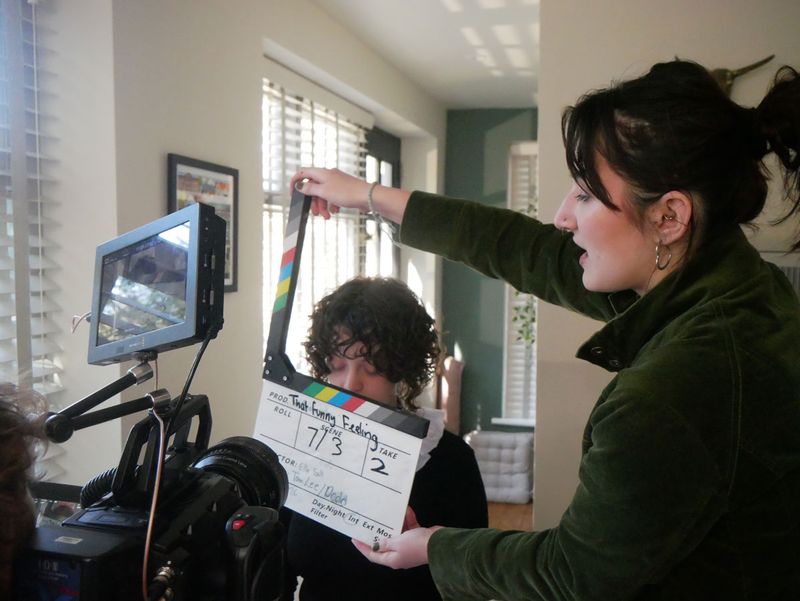 A person holds a film slate in front of a camera, preparing for a scene shoot in a well-lit room.